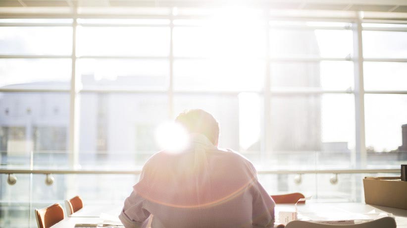 man sitting at a desk facing a wall of windows with a sun flare in the photo