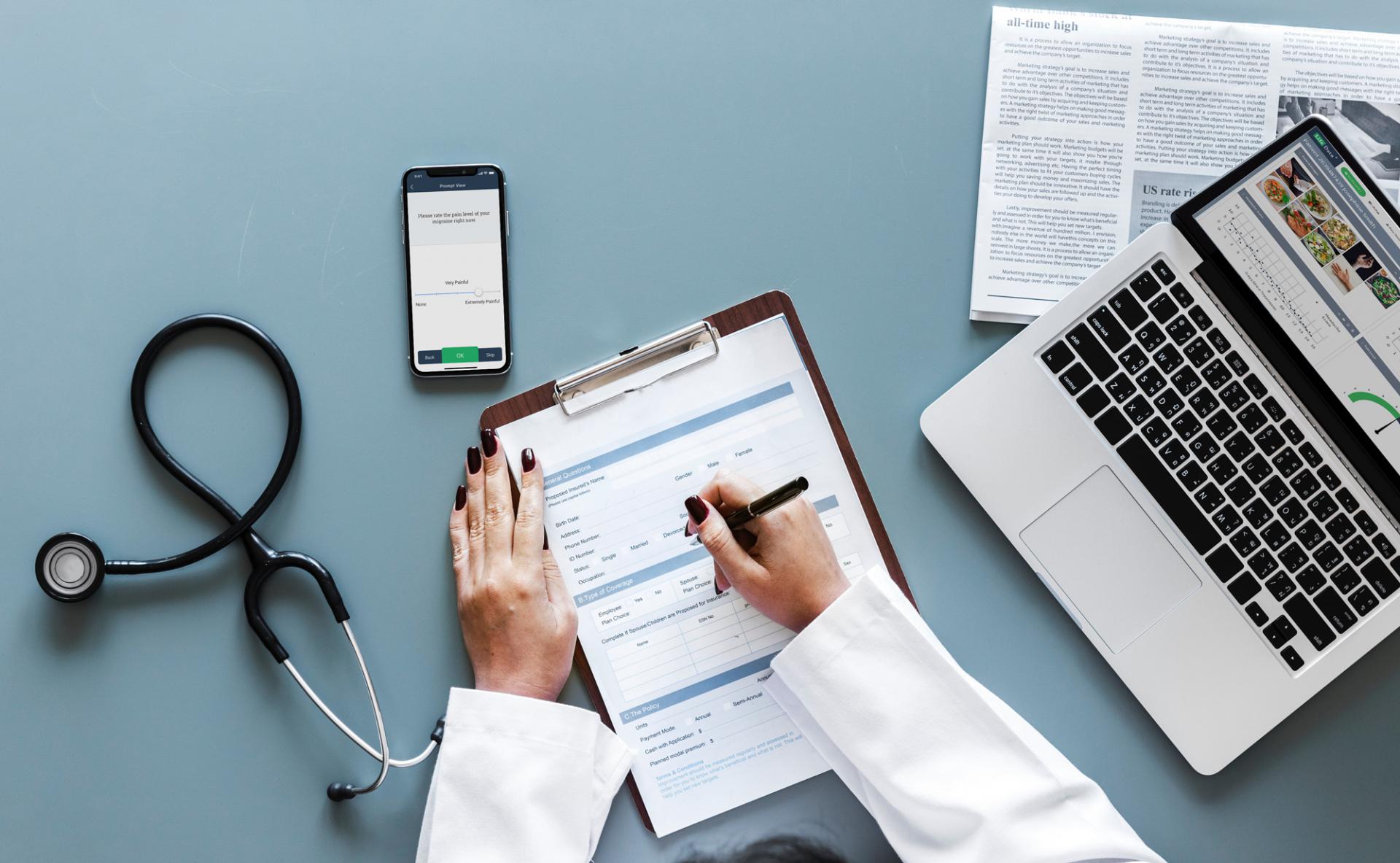 doctor writing on clipboard with Apple Macbook, smartphone, and stethoscope.